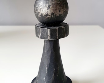 May include: A handcrafted, black metal pawn chess piece. The piece features a textured, cone-shaped body, a circular base, a hexagonal midsection, and a spherical top. The metal has a distressed, aged appearance with silver highlights.