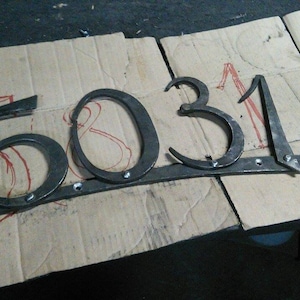 May include: Hand-forged metal house numbers, spelling out "5031". The numbers are dark gray and mounted on a horizontal bar. The numbers are on a cardboard surface with red markings and a marker.
