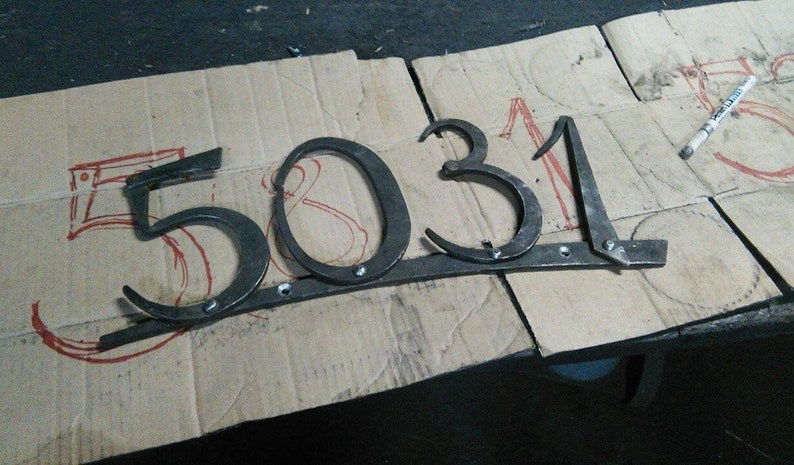 May include: Hand-forged metal house numbers, spelling out "5031". The numbers are dark gray and mounted on a horizontal bar. The numbers are on a cardboard surface with red markings and a marker.