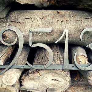 May include: Metal house numbers 9513 in a rustic style. The numbers are in a dark gray color and mounted on a horizontal bar. The background is a stack of wooden logs.