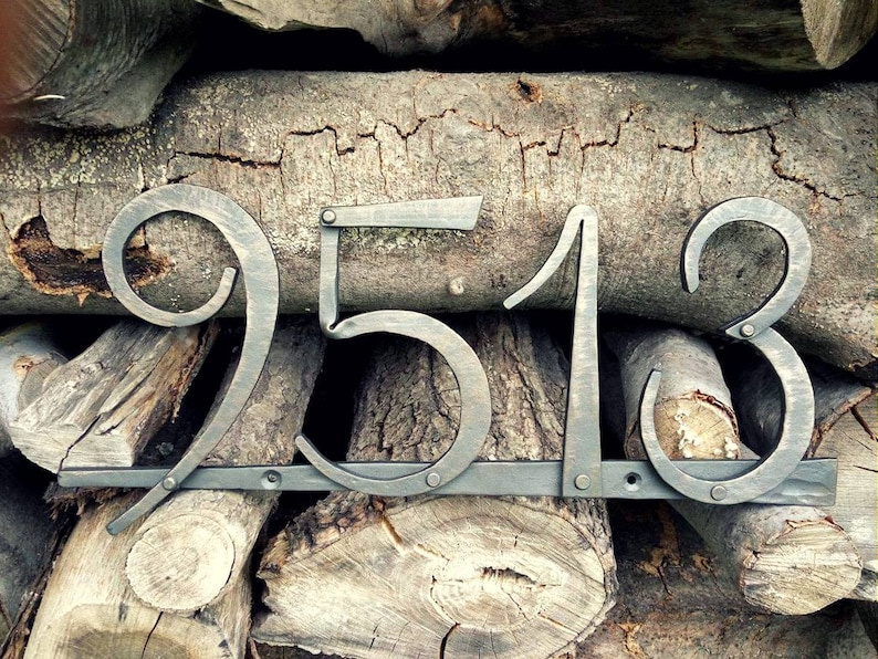 May include: Metal house numbers 9513 in a rustic style. The numbers are in a dark gray color and mounted on a horizontal bar. The background is a stack of wooden logs.