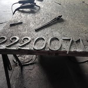 May include: Metal house numbers spelling out "22200711" are arranged on a wooden surface. The numbers are in a simple, sans-serif font and appear to be made of dark metal. Tools and other metal pieces are scattered around the numbers.