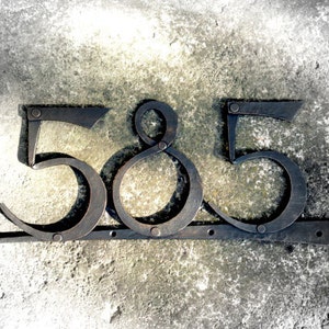 May include: Dark metal house numbers, "585", are mounted on a horizontal bar. The numbers are in a stylized font, with a rustic, industrial aesthetic. The numbers are attached with rivets.