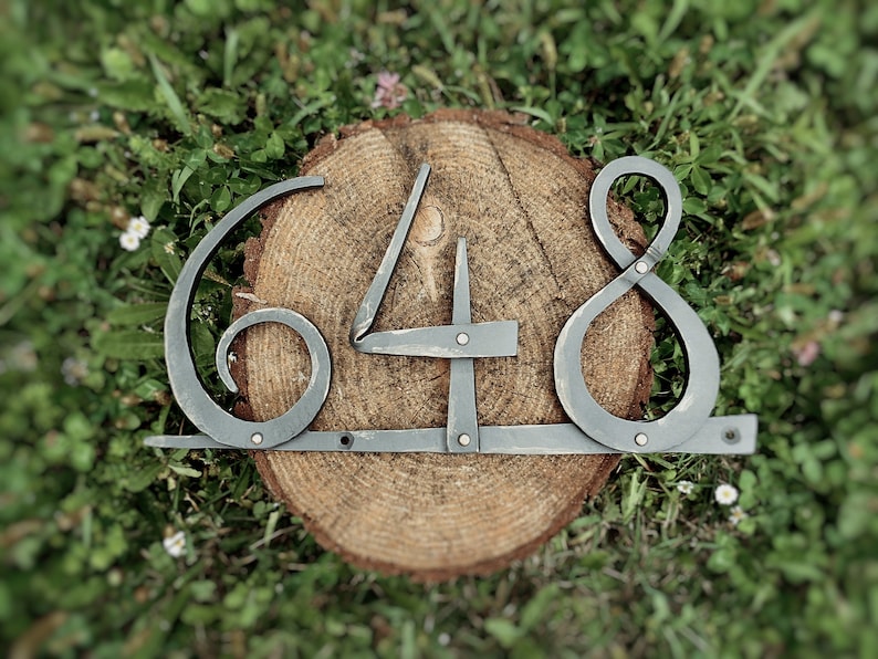 May include: Hand-forged metal house numbers, featuring the numbers 648, mounted on a wooden tree slice. The numbers are a dark gray color and have a rustic, handcrafted appearance. The background is green grass.