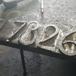 May include: Forged metal house numbers 17326, displayed on a textured surface. The numbers are in a simple, classic font, and appear to be freshly made. The metal has a dark, industrial finish.