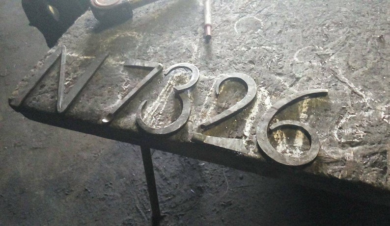 May include: Forged metal house numbers 17326, displayed on a textured surface. The numbers are in a simple, classic font, and appear to be freshly made. The metal has a dark, industrial finish.