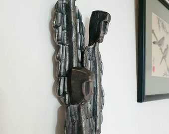 May include: A metal sculpture of three figures standing together. The figures are made of stacked metal pieces and have a rough, textured surface. The sculpture is painted in a dark gray color.