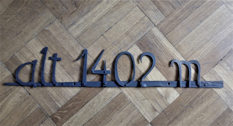 May include: A dark metal sign with the text "alt 1402 m" displayed on a wooden floor. The numbers and letters are crafted in a stylized font, with the "m" at the end. The sign is attached to a wooden base.
