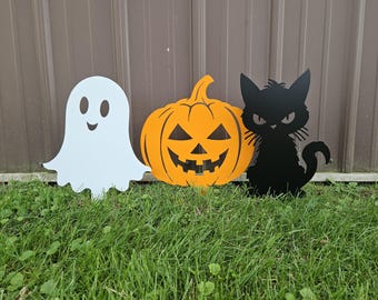 May include: Three Halloween yard decorations: a white ghost, an orange pumpkin with a jack-o'-lantern face, and a black cat.