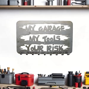 May include: A metal sign with the text "My Garage My Tools Your Risk" hanging on a white wall. The sign has a distressed look and is decorated with a brushed metal finish. The sign has several hooks on the bottom for hanging tools.