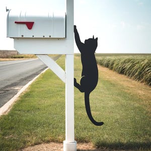 May include: A white mailbox with a red flag is mounted on a white post. A black metal cat silhouette is attached to the post, appearing to climb up the post.