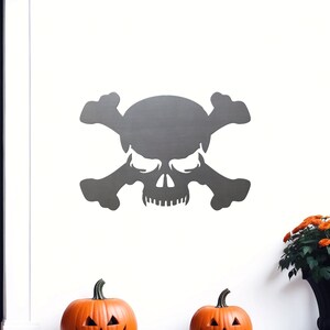 May include: A gray skull and crossbones silhouette on a white wall with two orange pumpkins in the foreground.