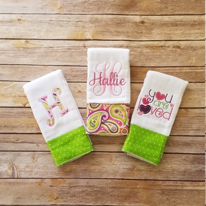May include: Three white burp cloths with green and pink trim. One cloth has the name "Hallie" embroidered on it, another has a large "H" embroidered on it, and the third has the words "you are loved" embroidered on it.