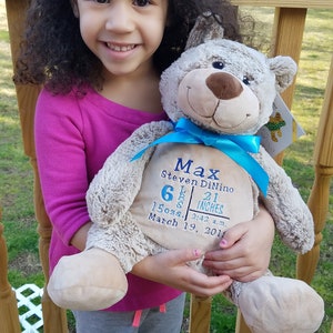 May include: A light brown teddy bear with a blue ribbon and embroidered details. The bear is holding a blue and white card with the text "Max Steven DiNino 6 lbs 21 inches 15 ozs. 2:43 a.m. March 19, 2011".
