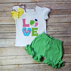 May include: White short-sleeved shirt with the word "LOVE" in colourful embroidered letters. The "O" is a beach ball and the "V" is a pair of flip flops. The shirt is paired with green shorts.