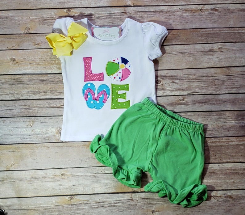May include: White short-sleeved shirt with the word "LOVE" in colourful embroidered letters. The "O" is a beach ball and the "V" is a pair of flip flops. The shirt is paired with green shorts.