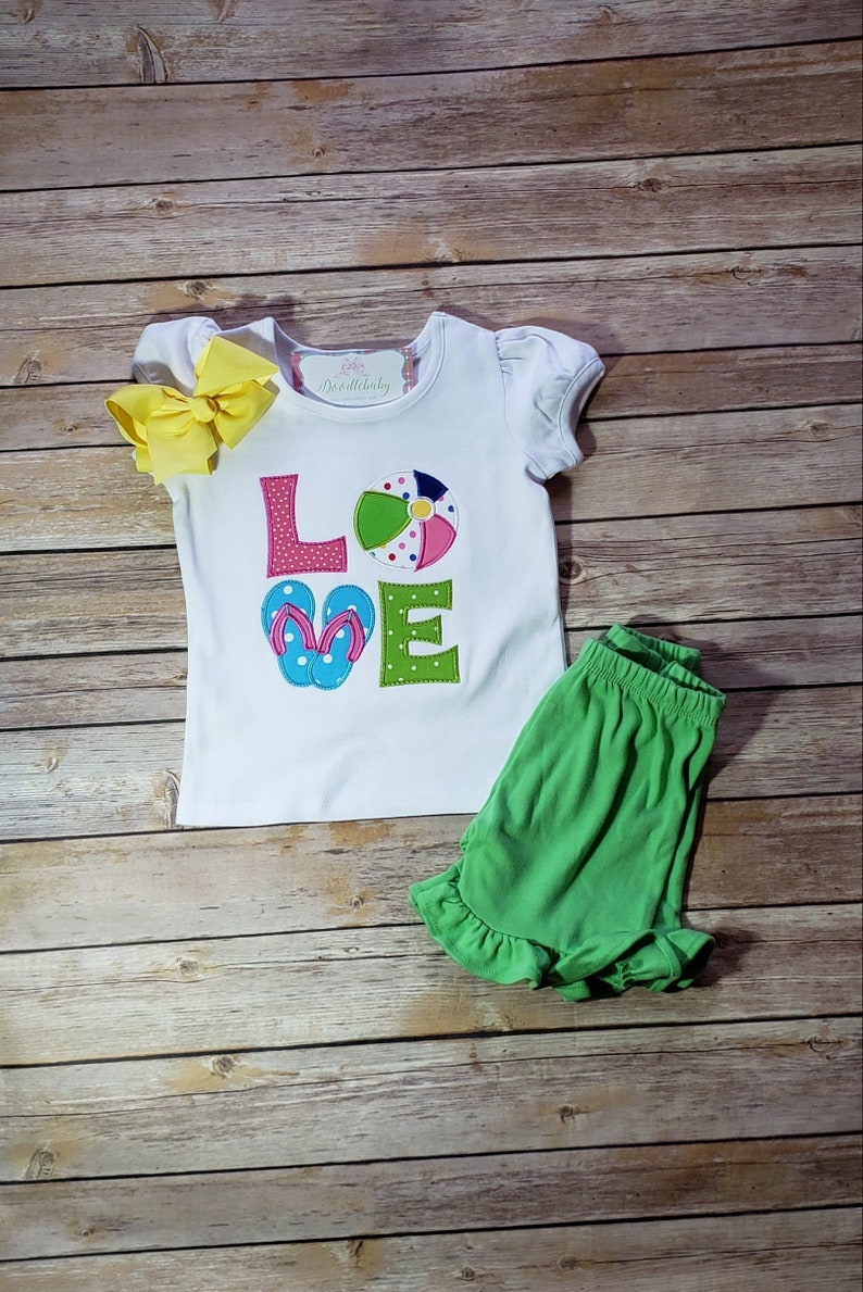May include: White t-shirt with the word "LOVE" spelled out in colourful felt letters. The "O" is a beach ball and the "V" is a pair of flip flops. The shirt is paired with green shorts.