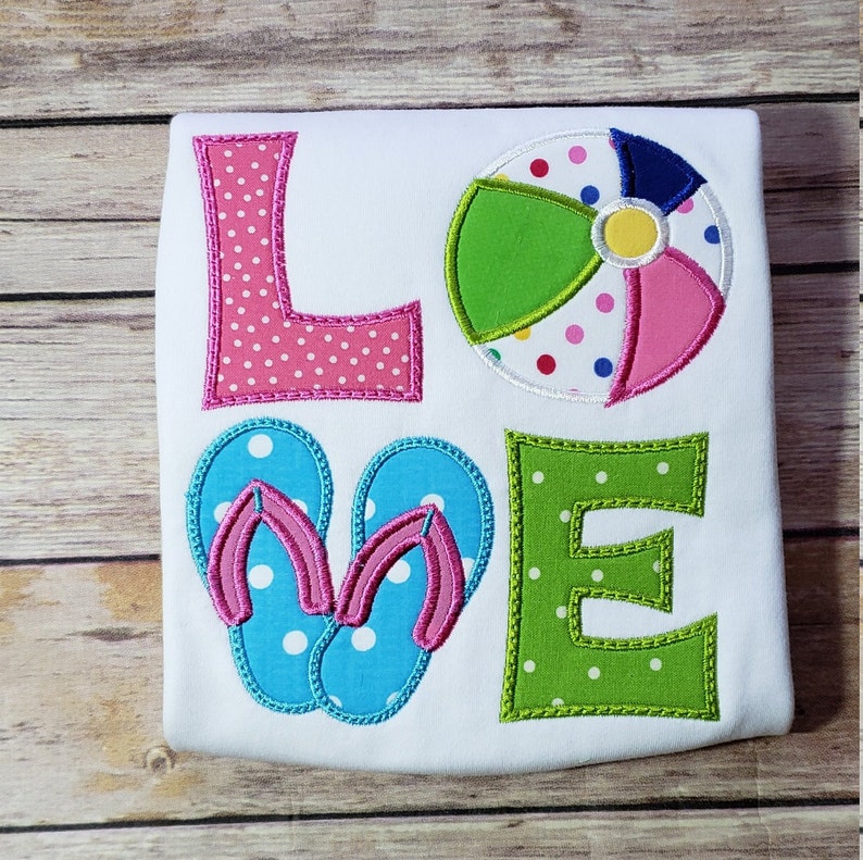 May include: A white t-shirt with the word "LOVE" spelled out in colourful letters. The letters are decorated with white polka dots. The "O" is replaced with a beach ball with pink, green, yellow, and blue sections. The "V" is replaced with a pair of blue flip-flops with pink straps.