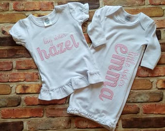 May include: Two white baby outfits with pink embroidery. The larger outfit is a short-sleeved t-shirt with the text "big sister hazel". The smaller outfit is a long-sleeved gown with the text "little sister emma".