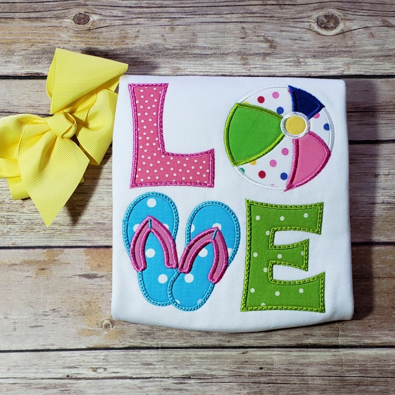 May include: White t-shirt with the word "LOVE" embroidered in colourful letters. The letters are decorated with polka dots. The "O" is replaced with a beach ball and the "V" is replaced with a pair of flip-flops.