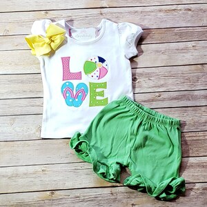 May include: White short-sleeved shirt with the word "LOVE" spelled out in colourful fabric appliques. The "O" is a beach ball and the "V" is a pair of flip flops. The shirt is paired with green shorts.