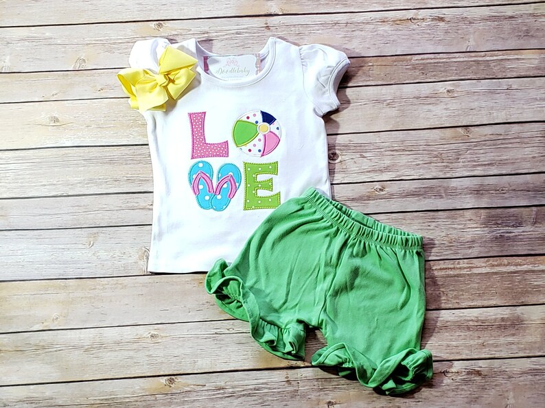 May include: White short-sleeved shirt with the word "LOVE" spelled out in colourful fabric appliques. The "O" is a beach ball and the "V" is a pair of flip flops. The shirt is paired with green shorts.