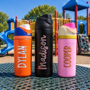 May include: Three cylindrical water bottles with flip-top lids. The left bottle is orange with the name DYLAN in silver. The center bottle is black with the name Madison in glittery pink. The right bottle is pink with the name COOPER in gold.