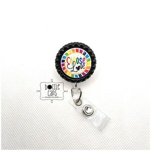 May include: A black bottle cap with a rainbow and glitter design. The cap has the words "Choose Love" written on it. The cap is attached to a clear retractable badge holder.