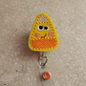 May include: A yellow candy corn-shaped badge holder with a smiling face, white eyes, and pink cheeks. The badge holder has a silver retractable reel and a clear plastic badge holder.