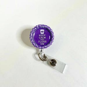 Könnte beinhalten: Lilafarbener und weißer Tupfen-Badgehalter mit einem runden lilafarbenen und weißen Badge, auf dem "Keep Calm and Floss On" steht, mit einer kleinen weißen Grafik eines Zahnseidebehälters.