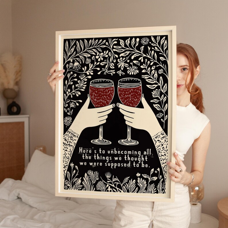 May include: A black and white print with a floral design and two wine glasses clinking. The text reads "Here's to unbecoming all the things we thought we were supposed to be."