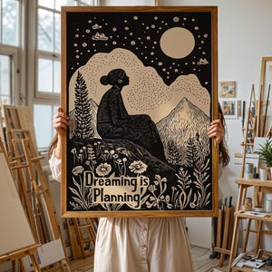 May include: A framed black and white print with a silhouette of a person sitting on a mountaintop with a moon and stars in the background. The print has the text "Dreaming is Planning" in gold lettering.