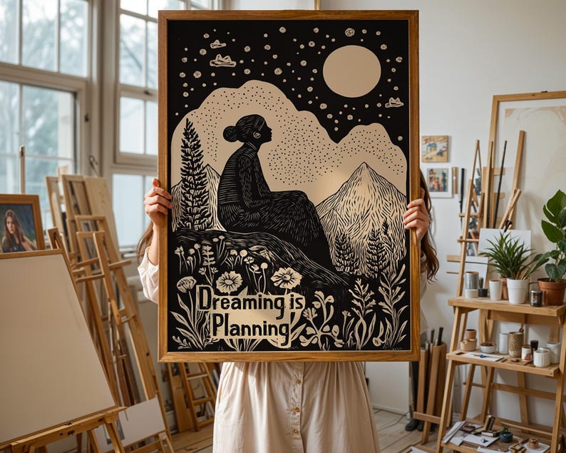 May include: A framed black and white print with a silhouette of a person sitting on a mountaintop with a moon and stars in the background. The print has the text "Dreaming is Planning" in gold lettering.