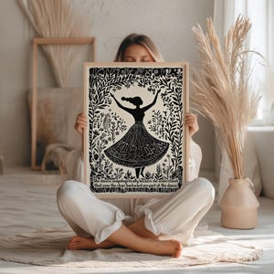 May include: Black and white art print featuring a silhouette of a dancing figure in a floral frame. The figure is wearing a long skirt. The print includes text at the bottom. The art is in a light wood frame.