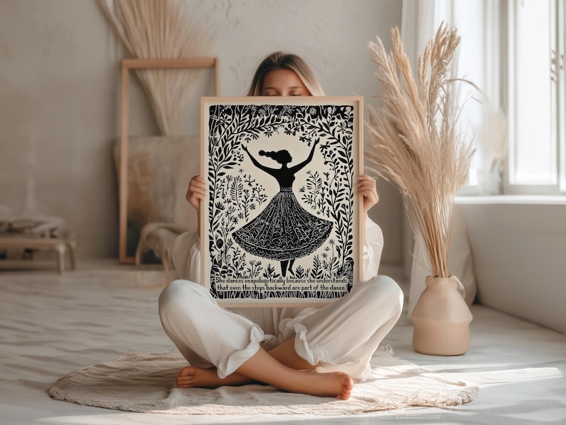 May include: Black and white art print featuring a silhouette of a dancing figure in a floral frame. The figure is wearing a long skirt. The print includes text at the bottom. The art is in a light wood frame.