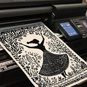 May include: A black and white print of a dancing woman surrounded by foliage is being printed on a large format printer. The print features a silhouette of a woman in a dress with her arms raised. The printer has a digital display.