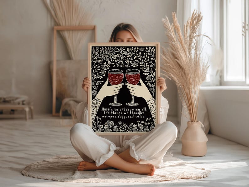 May include: Black and white illustration of two wine glasses with red wine, surrounded by floral designs. The text reads "Here's to unbecoming all the things we thought we were supposed to be."