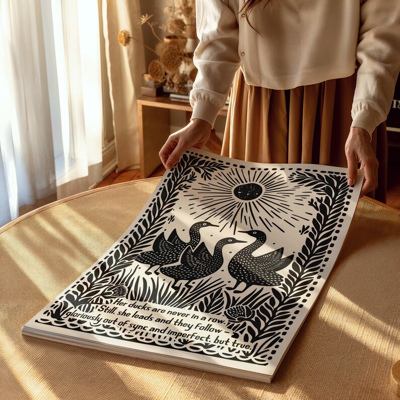 以下が含まれることがあります： 3羽の鳥が描かれた白黒のアートプリント。太陽の光線模様の下に配置され、装飾的な葉とテキスト「Her ducks are never in a row... gloriously out of sync and imperfect, but true.」が含まれています。