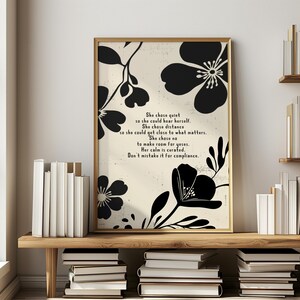 Puede incluir: Una impresi&oacute;n art&iacute;stica enmarcada con un borde dorado, que presenta dise&ntilde;os florales negros y texto inspirador. El texto dice: "She chose quiet...Don't mistake it for compliance". La impresi&oacute;n est&aacute; en un estante de madera con libros.
