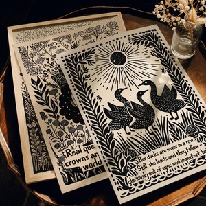 以下が含まれることがあります： 白黒のアートプリント。植物や動物のイラストが描かれています。1つのプリントには太陽の下に3羽のアヒルが描かれ、テキストが添えられています。別のプリントには花の模様とテキストがあります。プリントは木製のフレームに入っています。