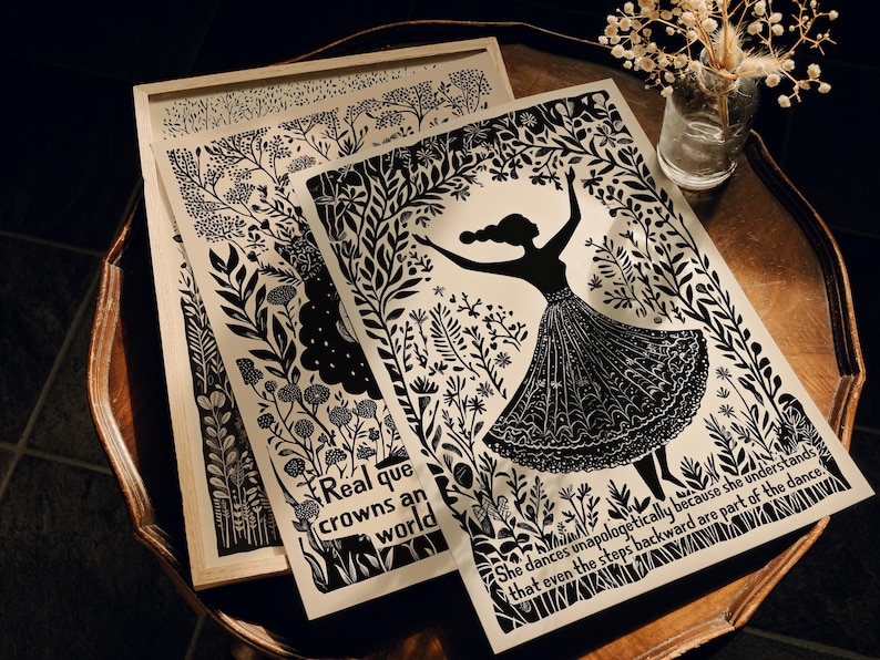 May include: Black and white art prints featuring botanical designs and a dancing figure. One print is framed in a light wood, and the others are layered on a wooden table. Text includes "Real que crowns an world" and a quote about dance.
