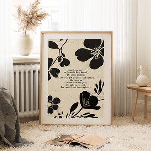 Puede incluir: Una impresi&oacute;n art&iacute;stica enmarcada con un fondo beige, motivos florales negros y texto. El texto dice "She chose quiet...Don't mistake it for compliance." La obra de arte se exhibe en una habitaci&oacute;n con un jarr&oacute;n de flores secas y un libro.