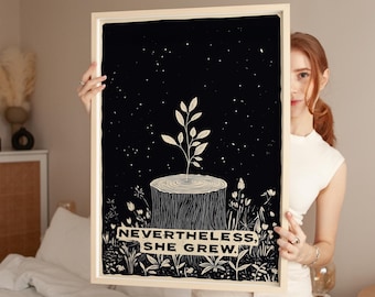 May include: A framed art print with a black background and a white illustration of a plant growing from a tree stump. The text "NEVERTHELESS, SHE GREW." is at the bottom. The frame is a light beige color.