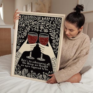 May include: A black and white print with a floral design. Two red wine glasses are raised in a toast. The text reads "Here's to unbecoming all the things we thought we were supposed to be."