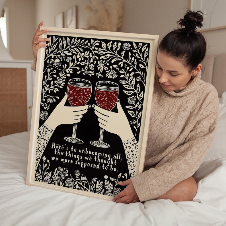 May include: A black and white print with a floral design. Two red wine glasses are raised in a toast. The text reads "Here's to unbecoming all the things we thought we were supposed to be."