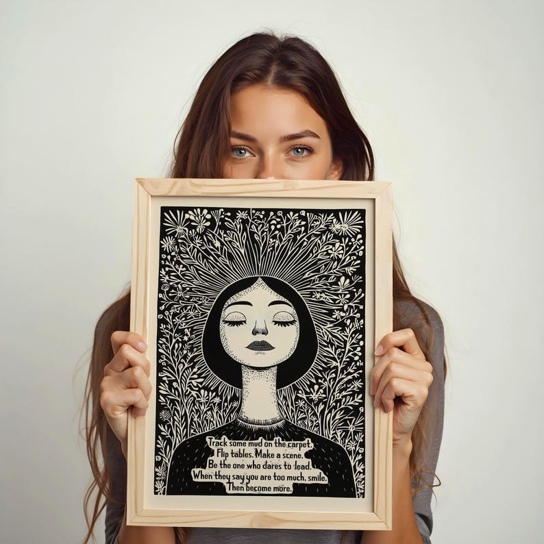 May include: Black and white illustration of a woman with closed eyes surrounded by flowers. The illustration is framed in a wooden frame. The text below the image reads: "Track some mud on the carpet. Flip tables. Make a scene. Be the one who dares to lead. When they say you are too much, smile. Then become more."