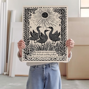 以下が含まれることがあります： 黒と白のアートプリント。3羽のアヒルが野原にいて、太陽と装飾的な縁取りがあります。プリントには「Her ducks are never in a row...gloriously out of sync and imperfect, but true.」というテキストが含まれています。