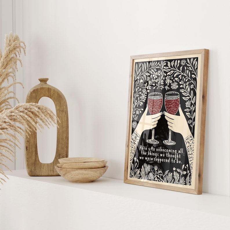 May include: A black and white illustration of two hands holding up wine glasses filled with red wine. The illustration is surrounded by a floral border and the text "Here's to unbecoming all the things we thought we were supposed to be."