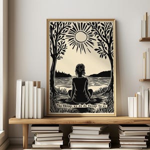 May include: A framed art print featuring a black and cream illustration of a person meditating by a lake under a sun. The artwork is framed in a gold-colored frame and includes the text, "The things we do do things to us."