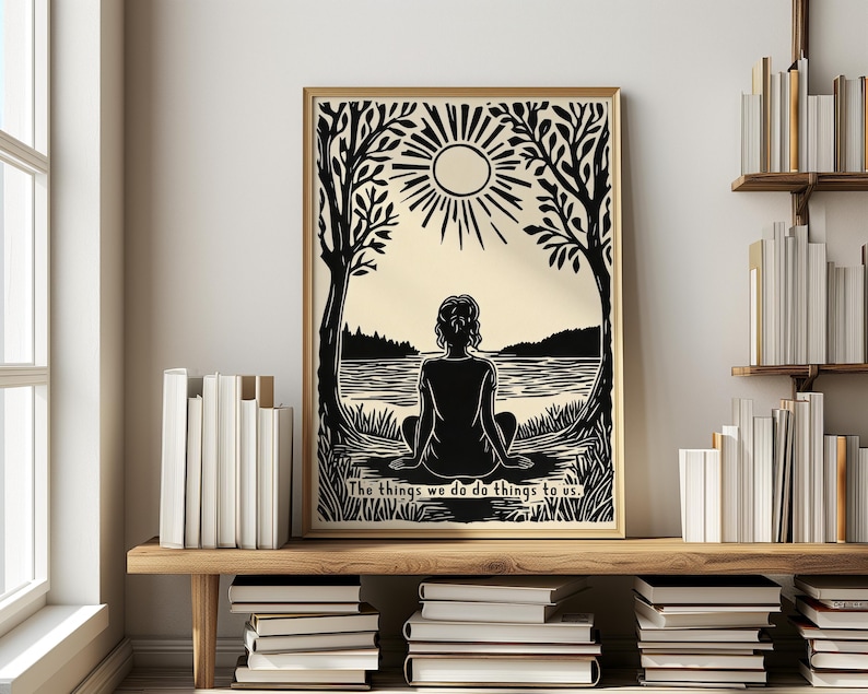 May include: A framed art print featuring a black and cream illustration of a person meditating by a lake under a sun. The artwork is framed in a gold-colored frame and includes the text, "The things we do do things to us."
